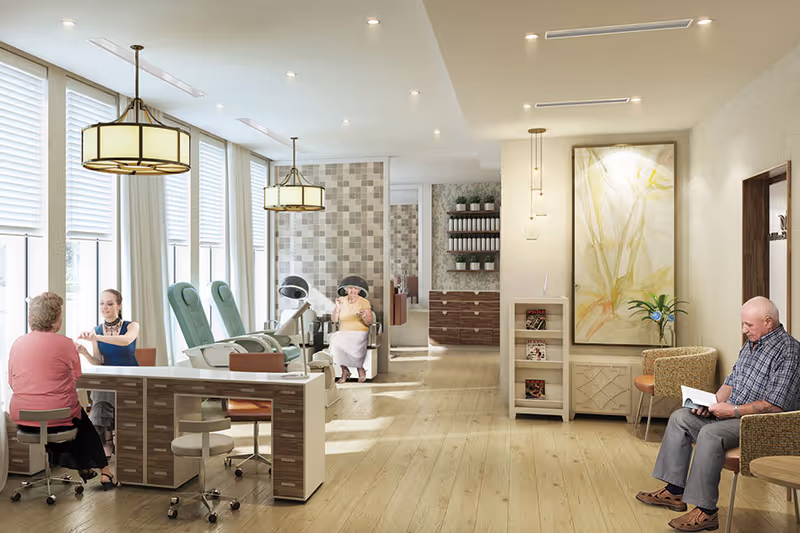 A bright and spacious senior care facility room with large windows letting in natural light. On the left, a woman is receiving a manicure from a staff member at a nail station with two green pedicure chairs behind them. In the background, an elderly woman wearing a hat is seated near a tiled wall. On the right side, an elderly man is sitting in a comfortable chair reading a book. The room has light wood flooring, modern pendant lights, a large abstract painting on the wall, and shelves with plants and magazines.