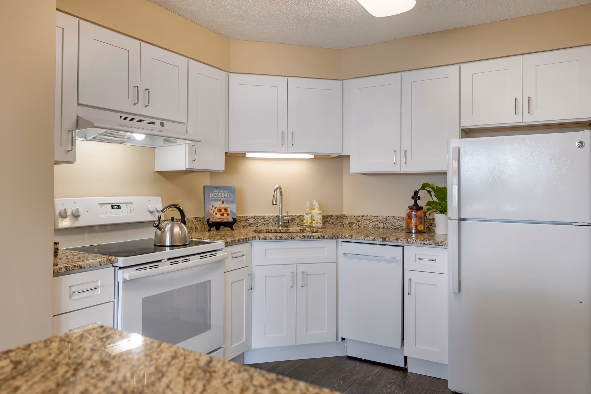 A modern kitchen with white cabinets, granite countertops, a white electric stove with a kettle on it, a dishwasher, a refrigerator, and a sink with a faucet. There is a cookbook titled 'Fine French Desserts' displayed on the counter, along with two soap dispensers and a decorative jar with fruit and a small plant.