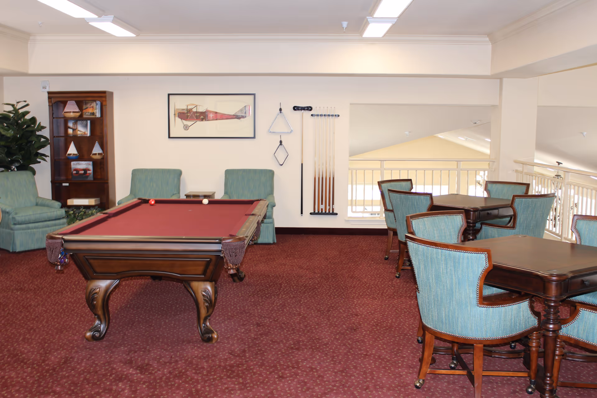 Interior view of a recreational room featuring a pool table with a red felt top, two green upholstered armchairs, a wooden corner shelf with decorative items, and several wooden tables with teal upholstered chairs. Pool cues and racks are mounted on the wall, and a framed picture of an airplane hangs above the armchairs. The room has a red carpet and cream-colored walls.