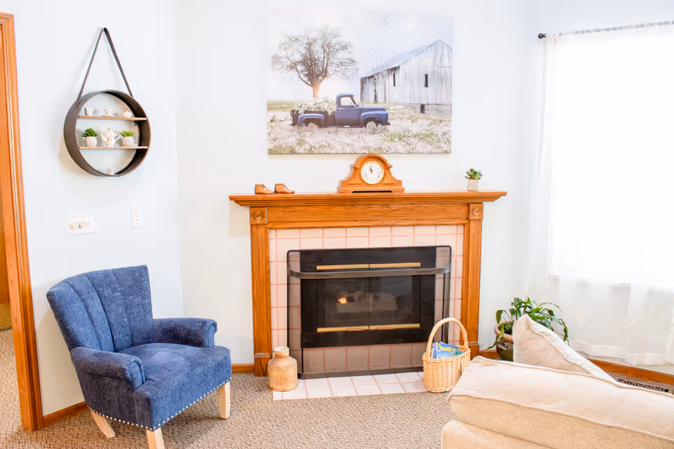 Cozy living room corner with a fireplace and wooden mantel, a blue armchair, beige sofa, and wall art above the mantel.