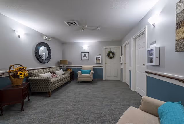 A well-lit interior hallway or common area in a senior living facility with a patterned sofa, two armchairs with blue cushions, a side table with a lamp, a basket of sunflowers, a round decorative mirror on the wall, framed artwork, and a door decorated with a wreath.