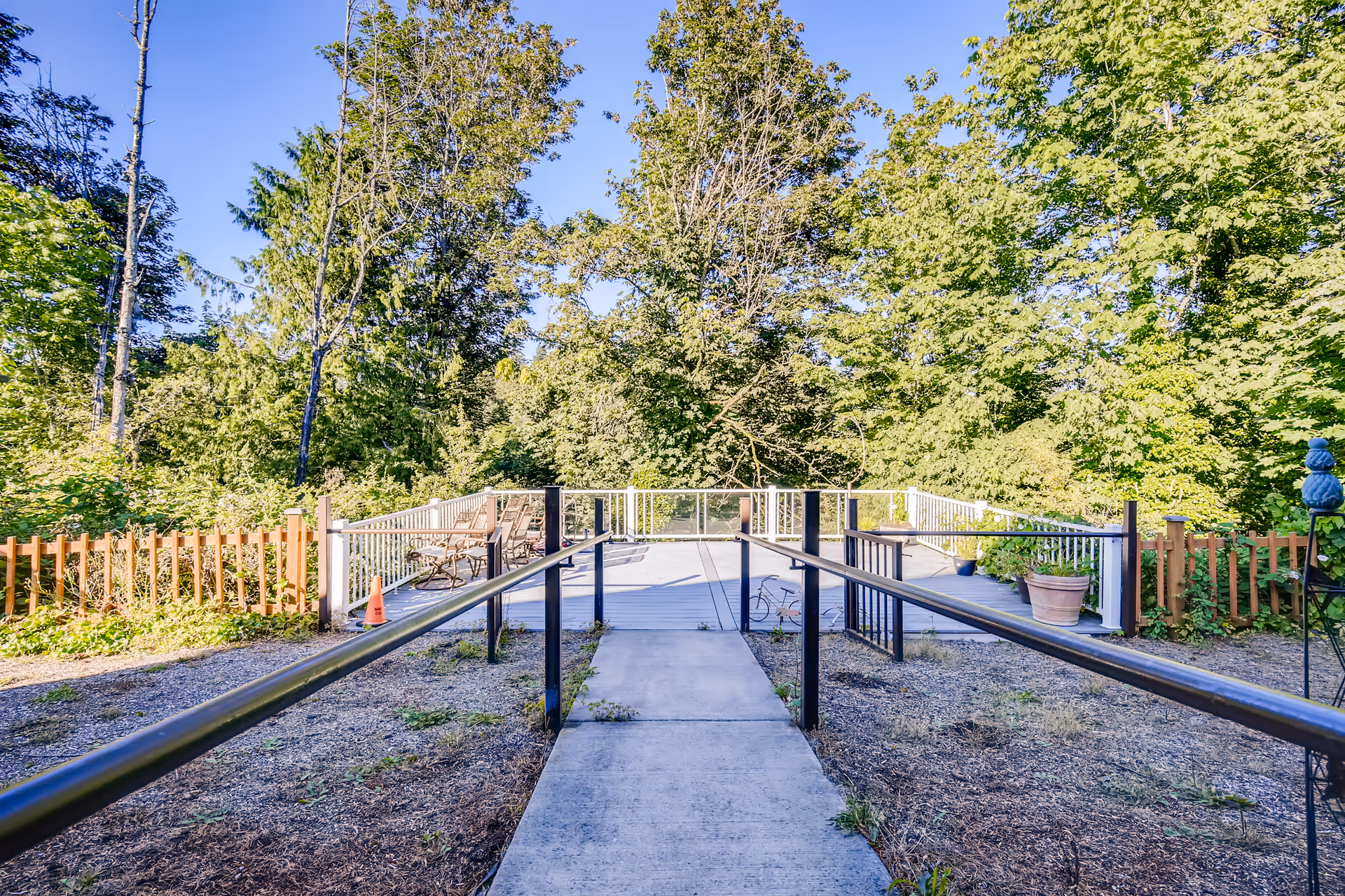 Concrete ramp with handrails leading to a wooden deck with chairs, surrounded by trees and greenery.