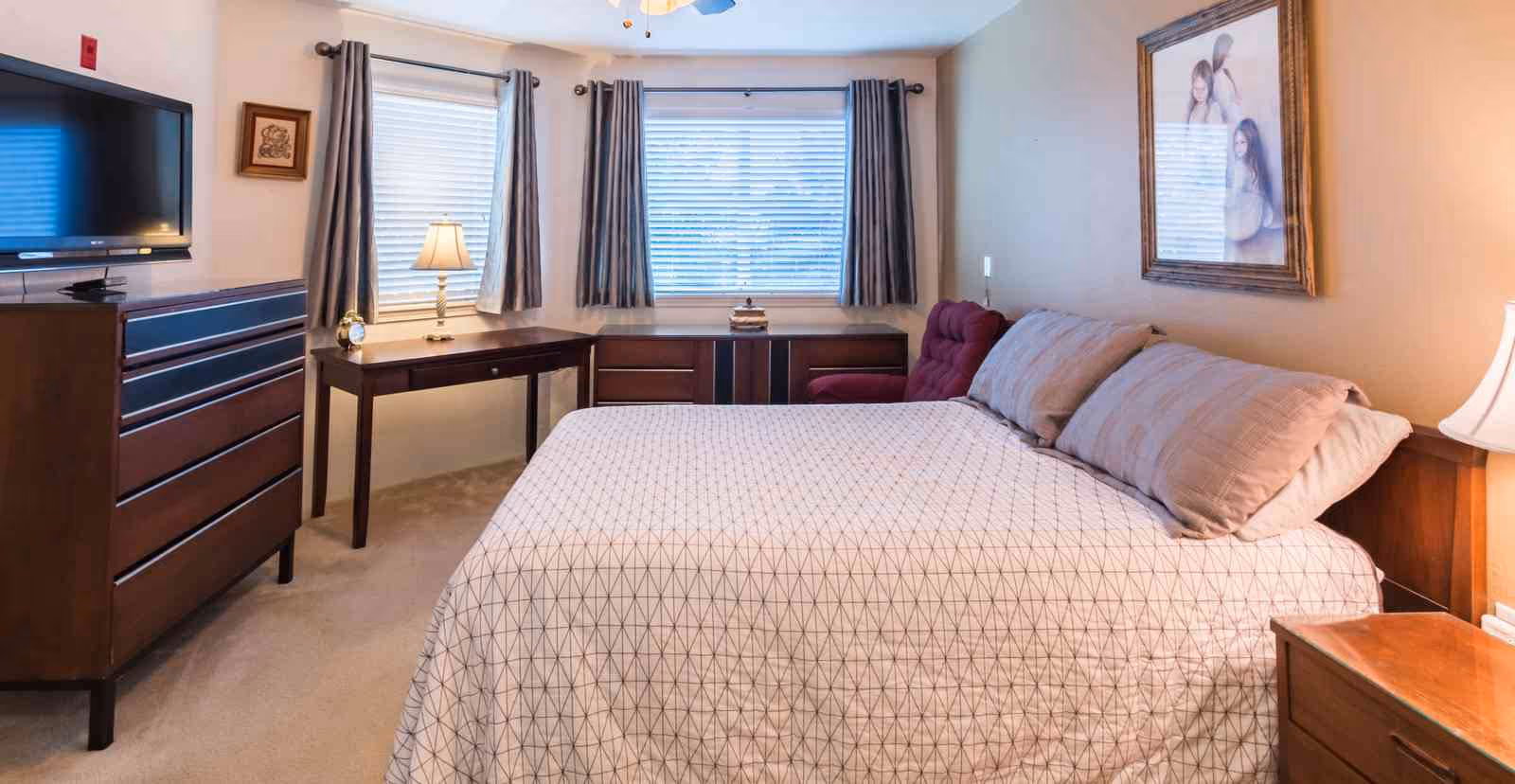 A bedroom with a large bed covered in a white and gray patterned bedspread, two pillows, a wooden dresser with a flat-screen TV on top, a small wooden desk with a lamp and clock, a window with gray curtains, a maroon armchair, and a framed picture on the wall.
