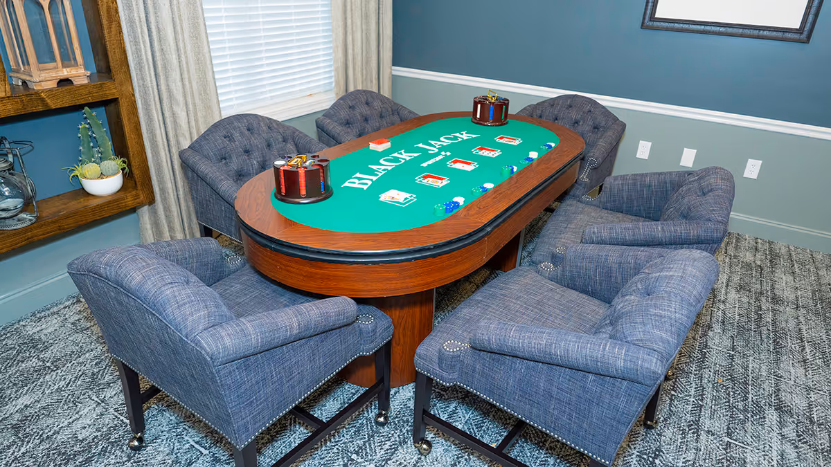 A blackjack gaming table with green felt surrounded by six upholstered blue-gray chairs in a carpeted room.
