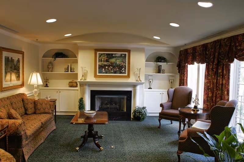 A cozy living room with a patterned sofa on the left, two upholstered armchairs on the right, a wooden coffee table in the center, and a fireplace with a framed painting above it. Built-in shelves flank the fireplace, decorated with plants and ornaments. Large windows with floral curtains allow natural light to enter the room.
