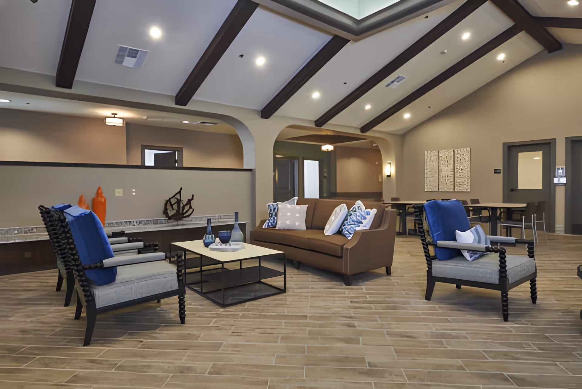 Bright, modern senior living lounge with a brown sofa, blue-accented armchairs, a coffee table, and a vaulted ceiling with exposed beams.