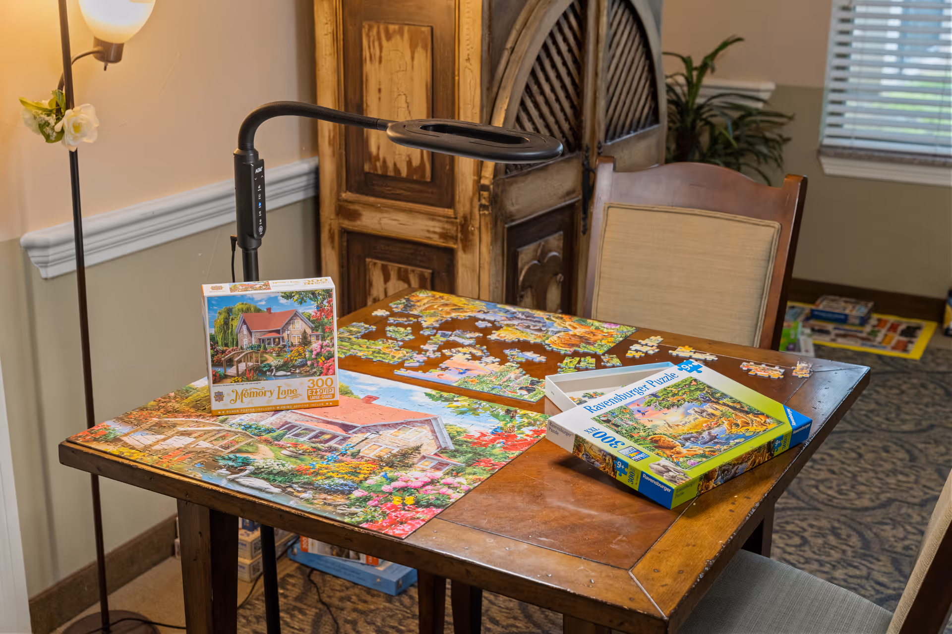A wooden table with two partially completed jigsaw puzzles and their boxes on top. The table is in a cozy room with a lamp, a wooden chair, a decorative cabinet, and a window with blinds. The floor is carpeted with a patterned rug.