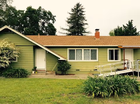 Single-story green house with a brown shingled roof, white door, and several windows. There is a wheelchair ramp on the right side of the house and green bushes and grass in the foreground. Tall trees are visible in the background under an overcast sky.