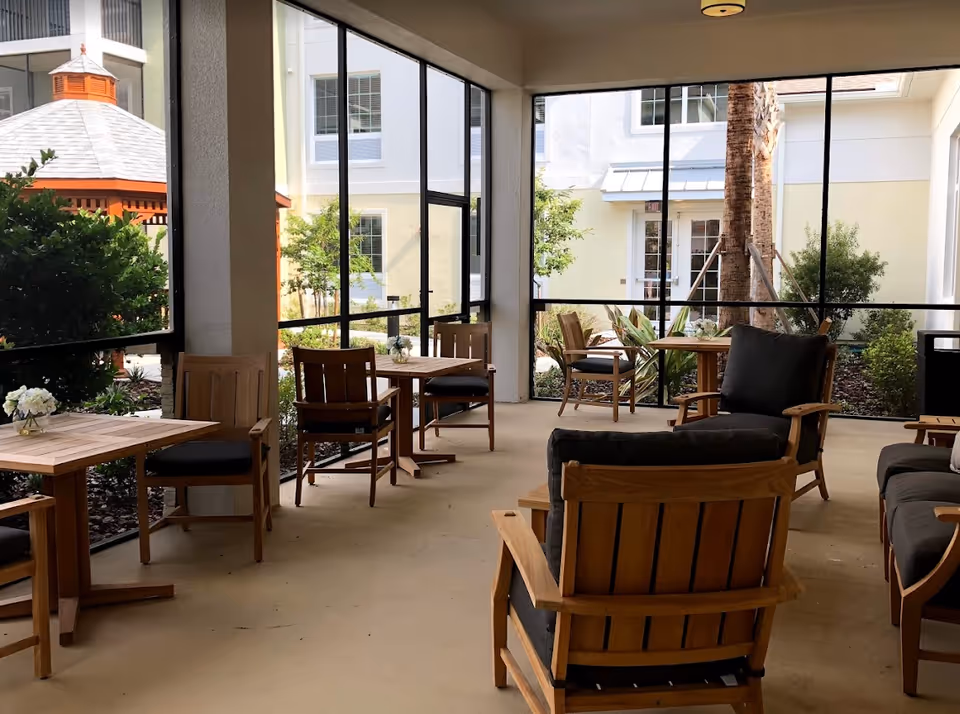 A screened-in patio area with wooden chairs and tables, some with black cushions, overlooking a garden with bushes, trees, and a gazebo. The patio is part of a building with white walls and windows.