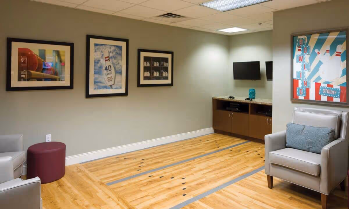 A cozy indoor recreational room with a wooden floor designed like a mini bowling lane, two gray armchairs with a blue cushion on one, a maroon ottoman, three framed pictures on the wall, a cabinet with a TV mounted above it, and a colorful bowling-themed poster on the right wall.