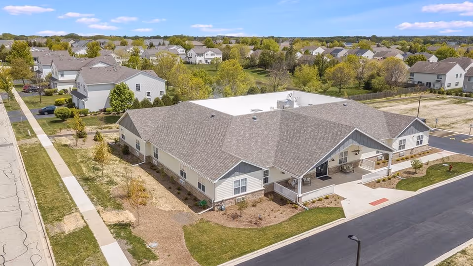 Aerial view of a single-story senior living building with gabled roof, covered front entrances, surrounding landscaping, and nearby houses.