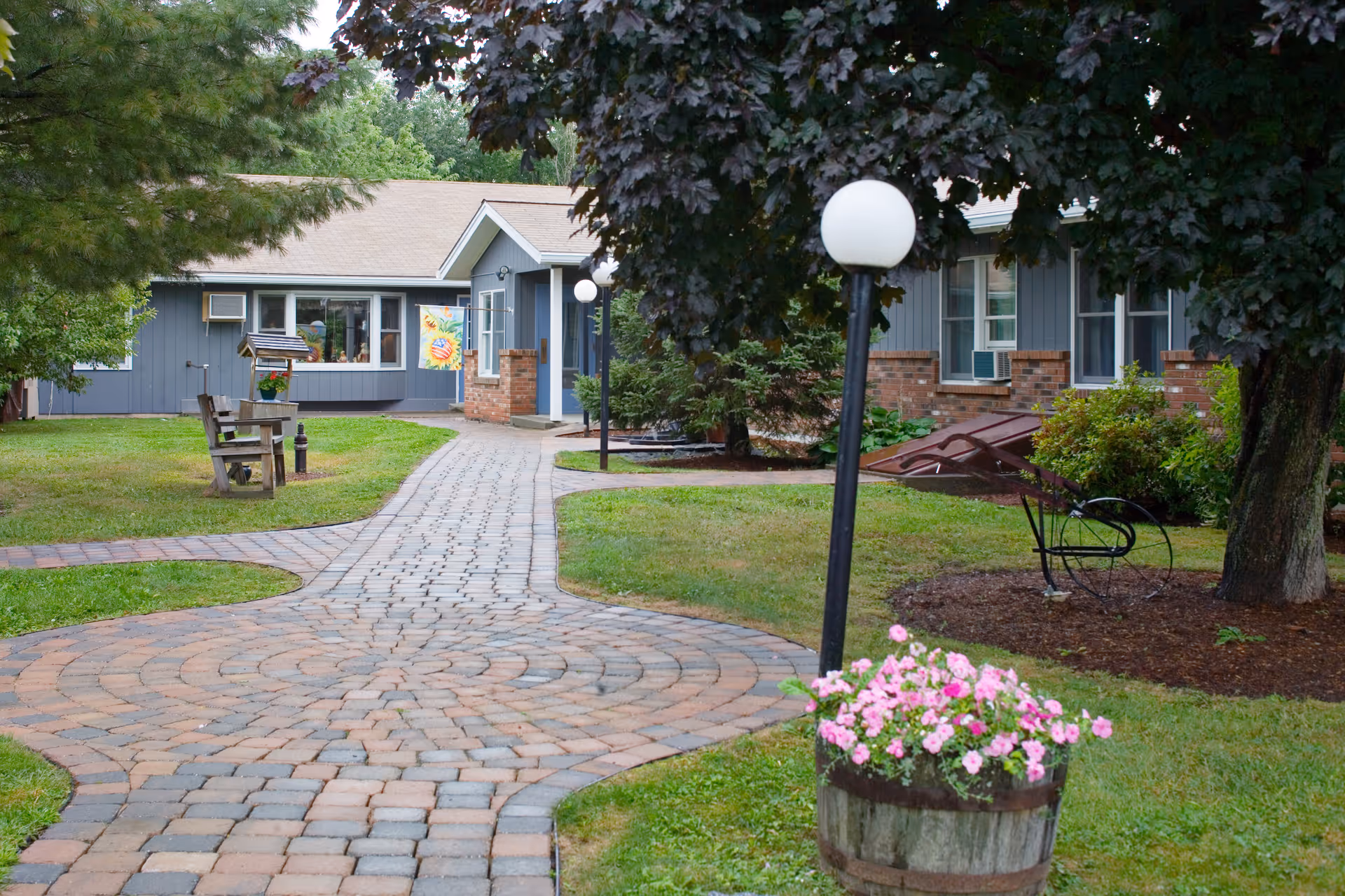 A paved walkway leads through a green lawn with trees and flower pots towards a single-story building with gray siding and brick accents. There are benches, lamp posts with round white lights, and a decorative wheelbarrow planter in the garden area.