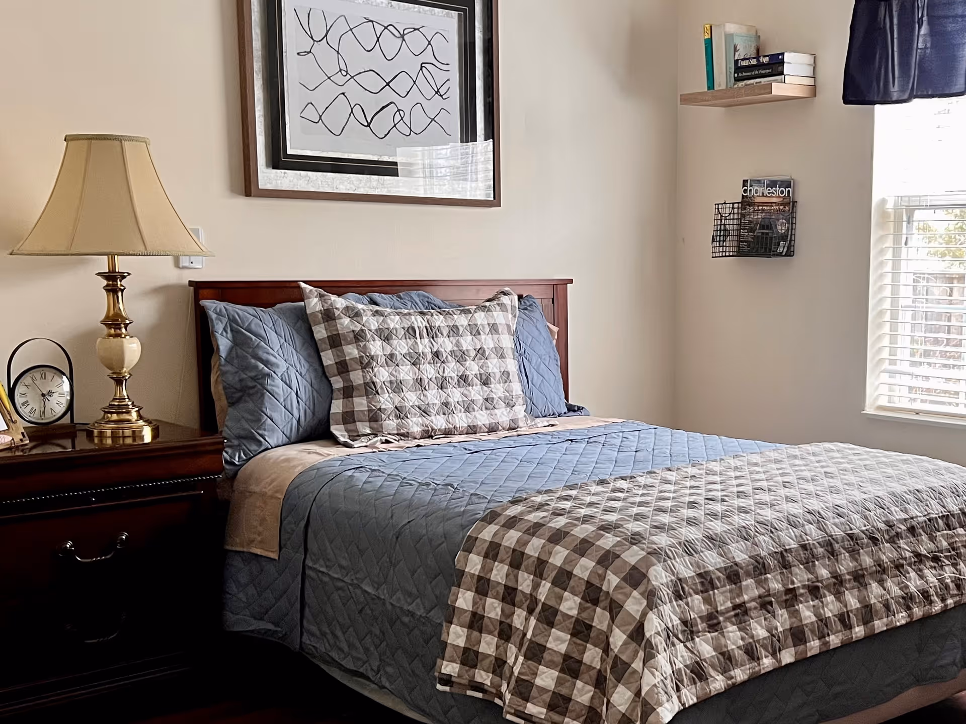 Sunlit bedroom with a made bed featuring a checkered quilt, a nightstand with a lamp and clock, framed art above, and a window to the right.