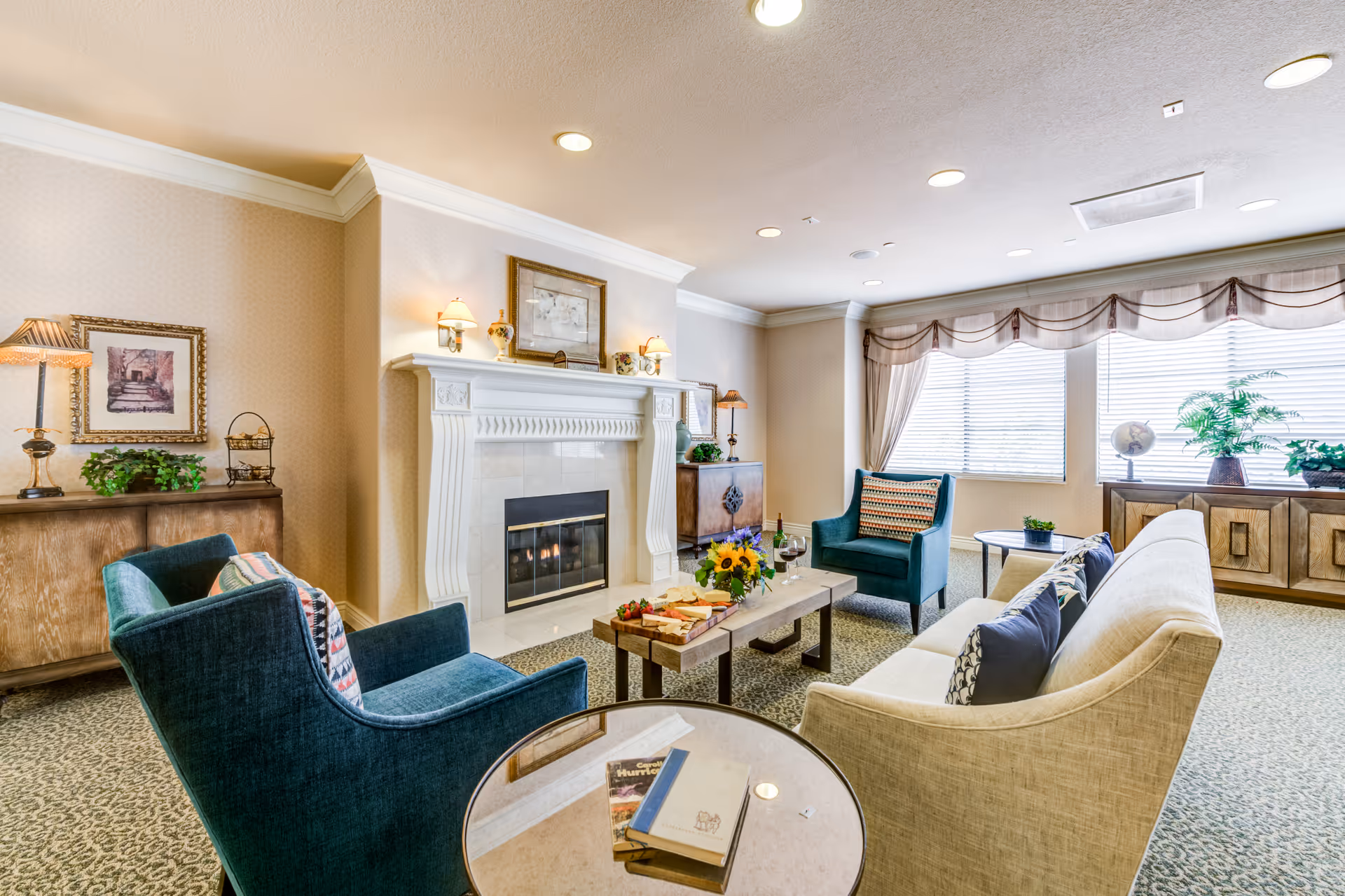 A cozy living room area in a senior living facility featuring a beige sofa with decorative pillows, two teal armchairs with patterned cushions, a wooden coffee table with a flower arrangement, wine glasses, and a cheese platter. The room has a white fireplace with wall sconces and framed artwork above it. Large windows with sheer curtains allow natural light to fill the room. There are plants and decorative items on wooden sideboards along the walls.