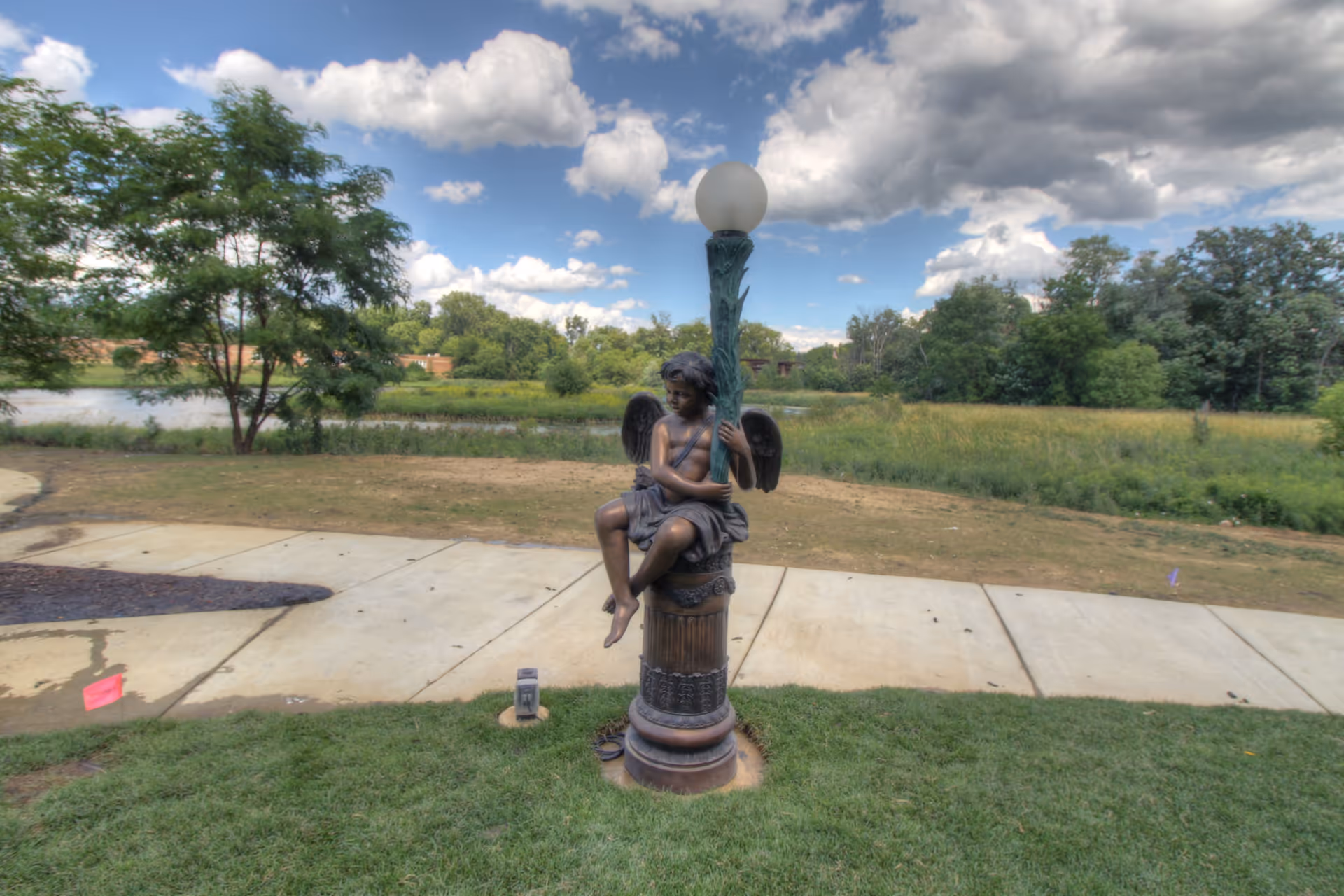Bronze angel statue holding a lamp post on a lawn by a sidewalk with trees and a pond in the background under a partly cloudy sky.