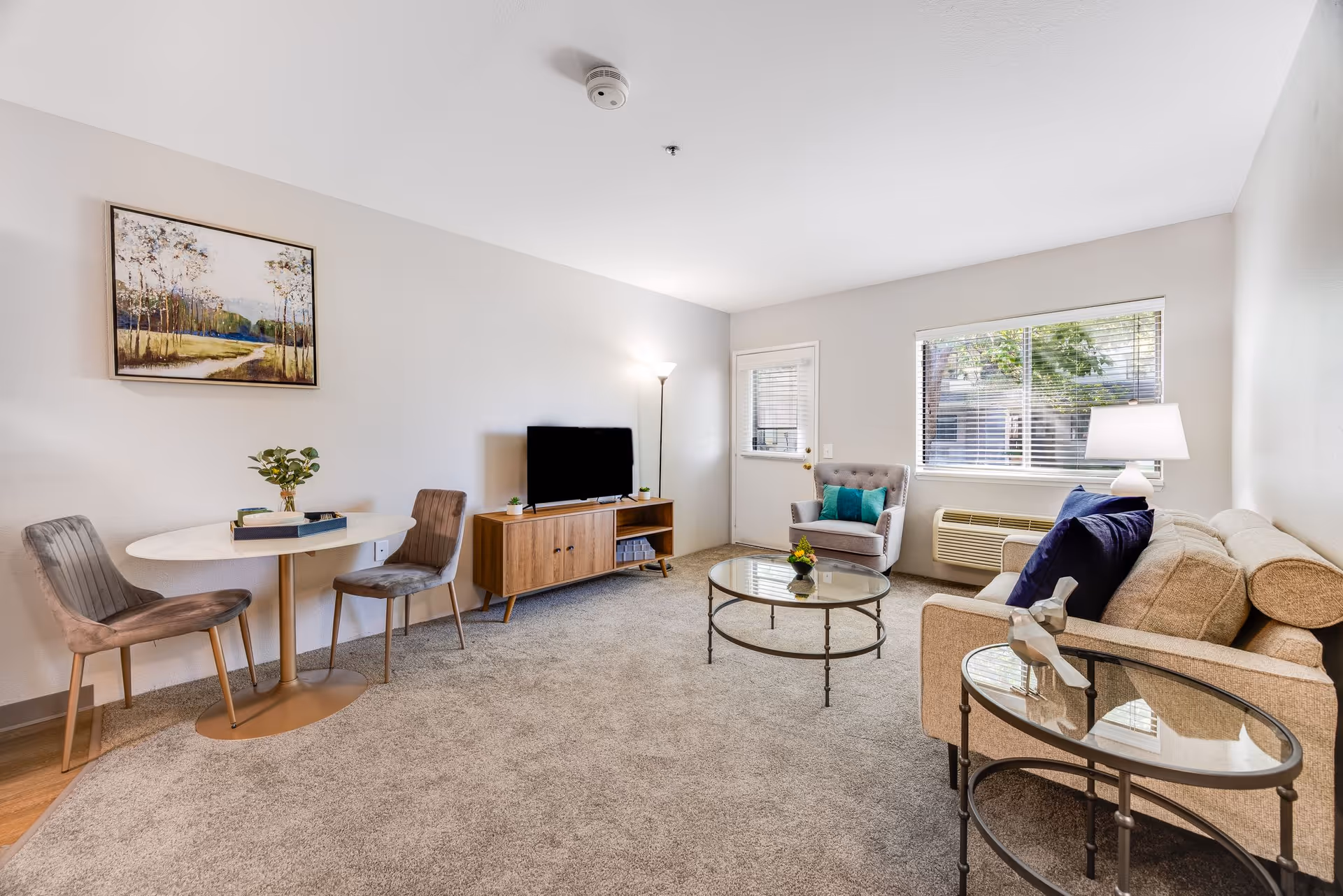 A bright living room with beige carpet and light-colored walls. The room features a beige sofa with dark blue pillows, a glass coffee table, a round side table with a decorative item, a gray armchair with a teal pillow, a wooden TV stand with a flat-screen TV, a floor lamp, and a small round dining table with two chairs. A large window with blinds and a door are visible in the background, along with a framed landscape painting on the wall.