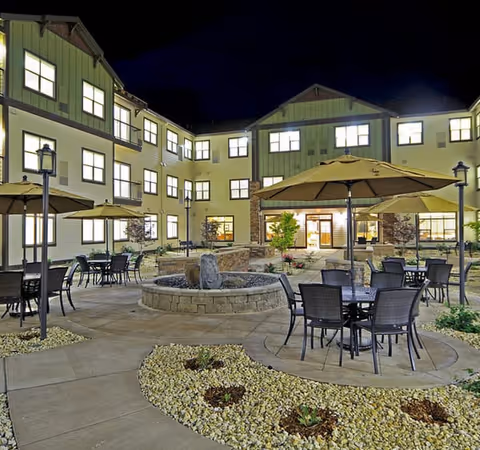 Night view of an outdoor courtyard area at a senior living facility with multiple tables and chairs under large umbrellas, surrounded by a three-story building with many lit windows. The courtyard features a circular stone water fountain and landscaped areas with rocks and small plants.