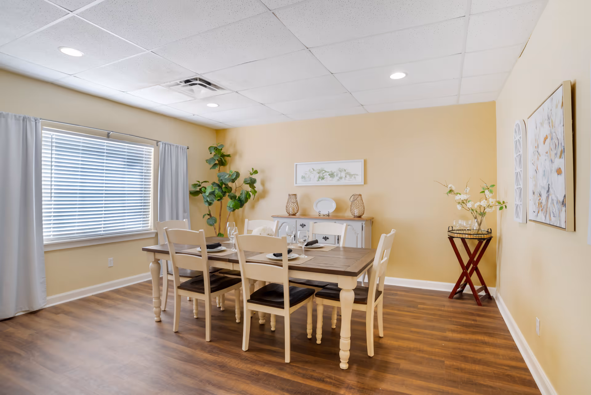 A dining room with a wooden table set for six people with plates, glasses, and napkins. The room has light yellow walls, a large window with white blinds and light gray curtains, a wooden sideboard with decorative items, a tall green plant in the corner, and a small table with a vase of flowers. The floor is wood, and the ceiling has recessed lighting.