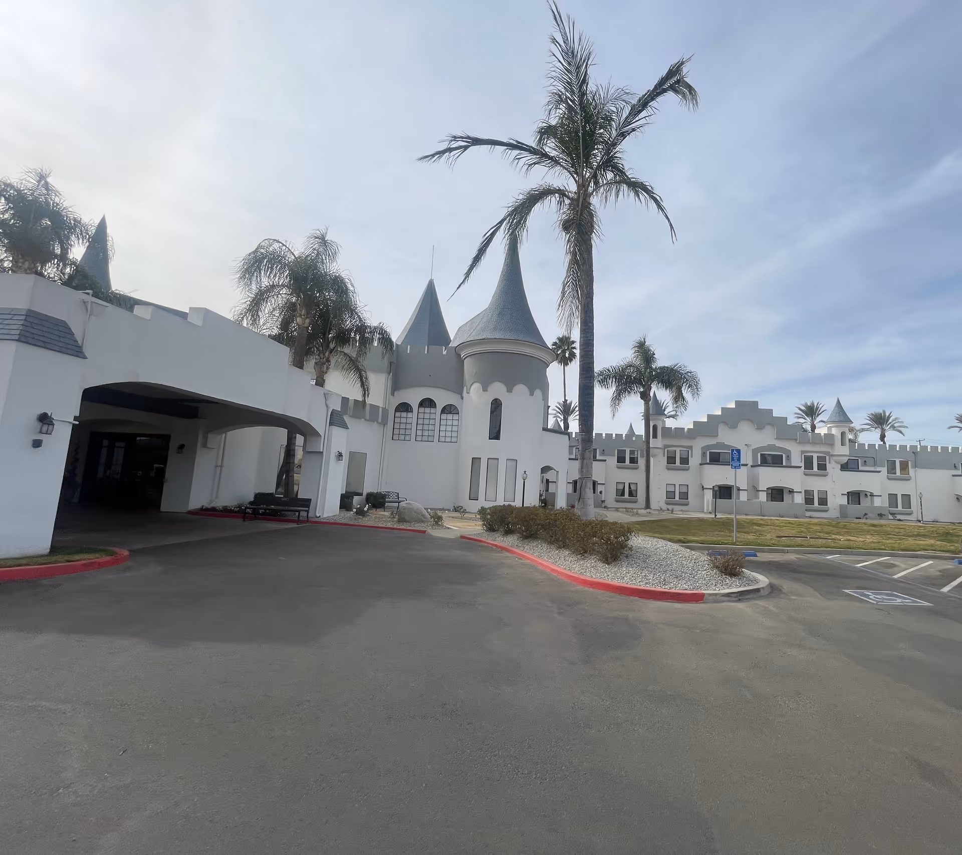 Exterior view of a senior living community building designed like a castle with white walls, turrets, and arched windows. Palm trees and a parking lot with handicap parking spaces are visible in front of the building under a partly cloudy sky.