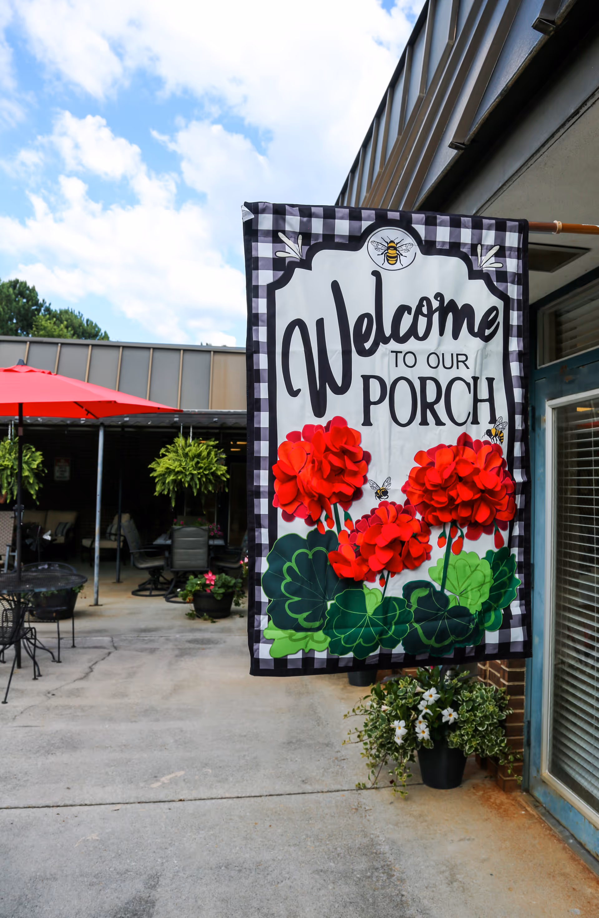 A colorful 'Welcome to Our Porch' flag with red flowers hangs outside a covered patio with tables and potted plants.