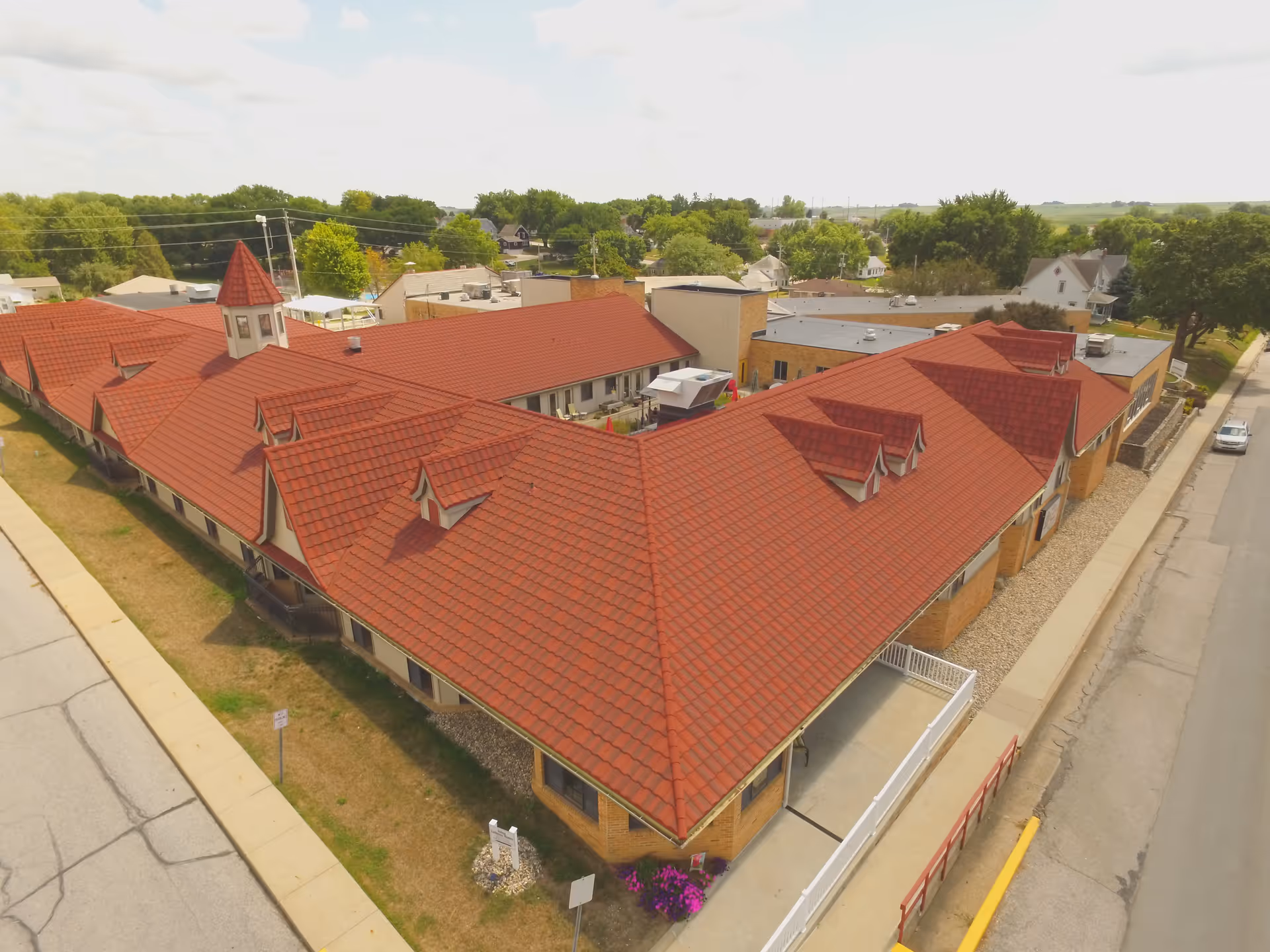 Aerial view of Salem Lutheran Homes and Rehabilitation Center showing a large building with a red tiled roof, multiple dormer windows, and a small tower. The building is surrounded by sidewalks, streets, and some greenery with trees and houses in the background.