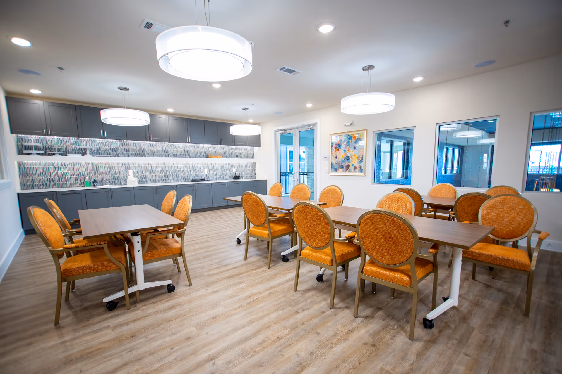A bright dining room with wooden floors, several tables with orange cushioned chairs, modern pendant lights hanging from the ceiling, and a long counter with cabinets and a decorative backsplash along one wall.