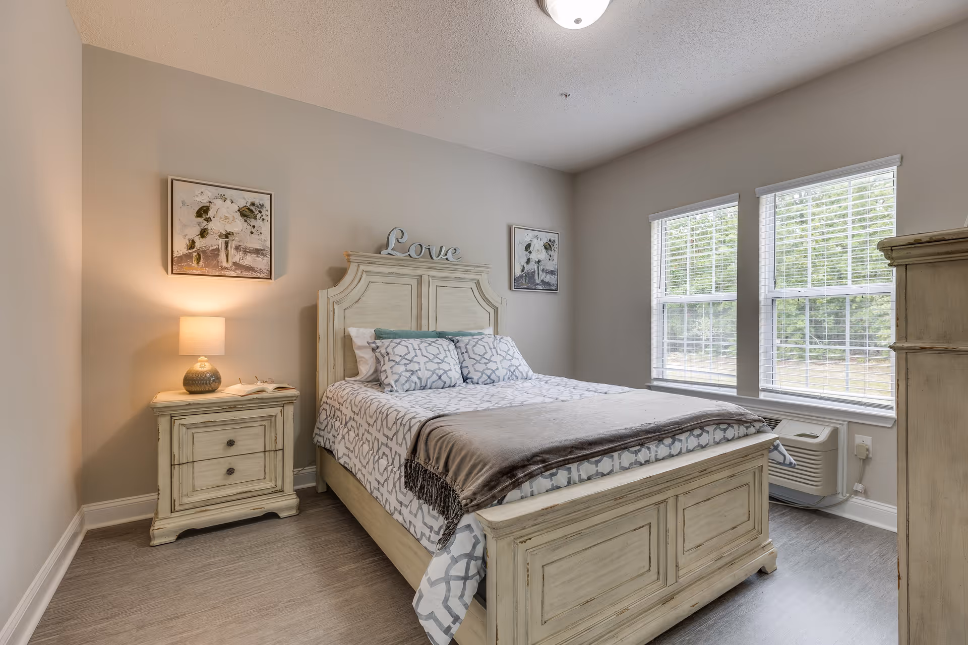 A cozy bedroom with a wooden bed frame and matching nightstand. The bed is made with patterned bedding and pillows, and a decorative 'Love' sign is placed on the headboard. There is a lamp on the nightstand, two framed floral paintings on the wall, and two large windows with blinds letting in natural light. The room has light-colored walls and flooring.