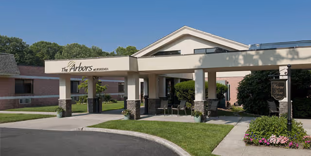 Covered entrance and driveway of The Arbors facility with columns, seating, and landscaped grounds.