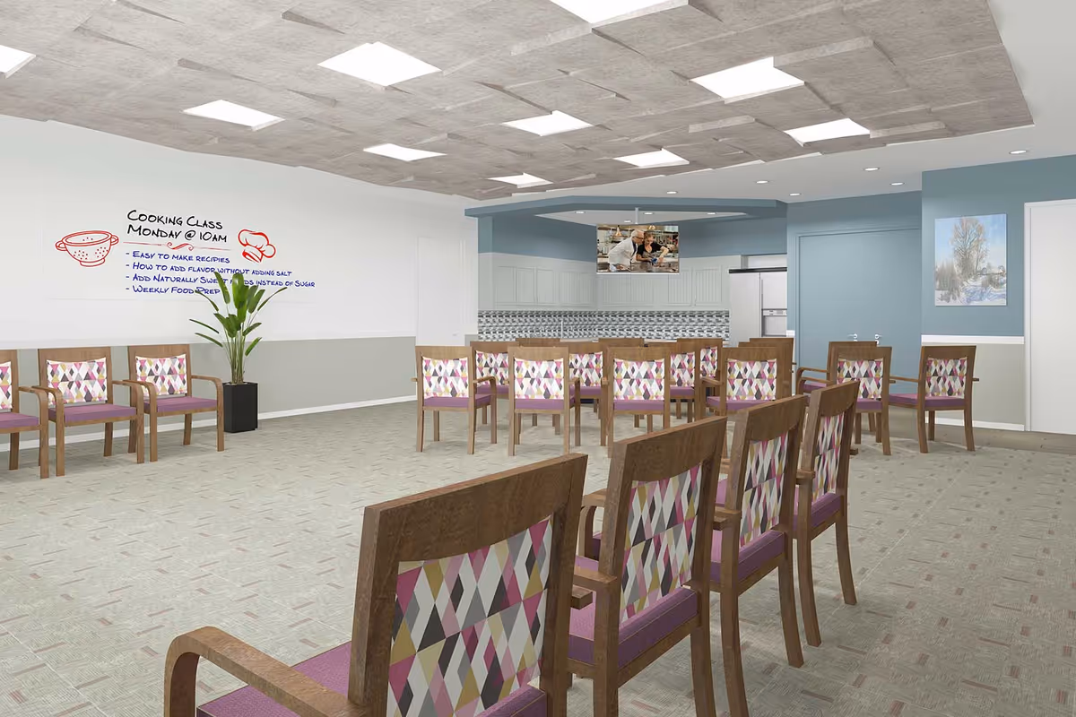 A spacious room with multiple wooden chairs featuring colorful patterned backs and purple seats arranged facing a kitchen area. The kitchen has white cabinets, a tiled backsplash, and a refrigerator. A flat-screen TV hangs from the ceiling showing two people cooking. On the left wall, there is a whiteboard with a drawing of a colander and text about a cooking class scheduled for Monday at 10 AM. A potted plant is placed below the whiteboard. The ceiling has recessed lighting and a modern design with square light panels.