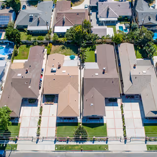 Aerial view of a residential neighborhood showing several single-story houses with driveways, lawns, and backyards. The houses are arranged in a neat row along a street with sidewalks and greenery.