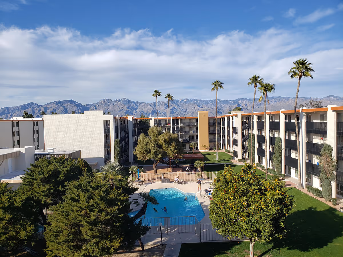 Outdoor view of Fellowship Square Tucson senior living facility with a swimming pool in the center, surrounded by green trees and grass. The multi-story building forms a U-shape around the pool area. Tall palm trees are scattered throughout the grounds, and mountains are visible in the background under a partly cloudy sky.