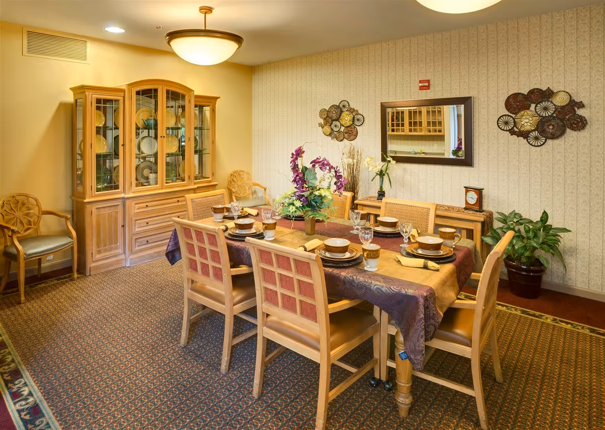 A dining room with a rectangular wooden table set for six people with matching chairs. The table is covered with a decorative tablecloth and has a floral centerpiece. Behind the table is a wooden china cabinet displaying plates and bowls. The room has patterned carpet, wallpapered walls with metal wall art, a mirror, a clock on a sideboard, and a potted plant in the corner.