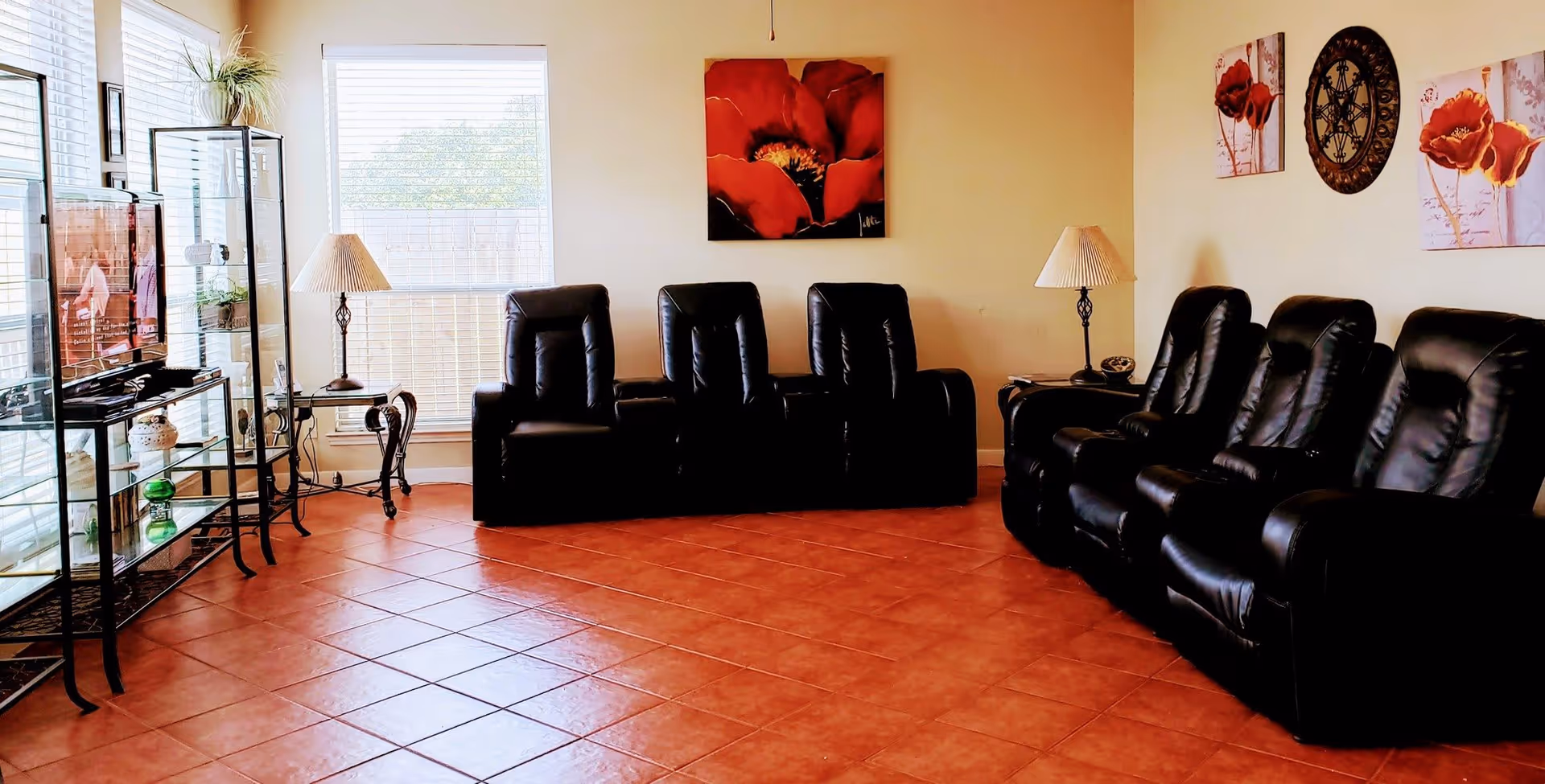 A living room with black leather recliner chairs arranged in an L-shape along two walls. The room has terracotta tiled flooring and cream-colored walls adorned with red floral paintings and a decorative wall piece. There are two windows with blinds letting in natural light, a TV on a glass stand, and two side tables with lamps.