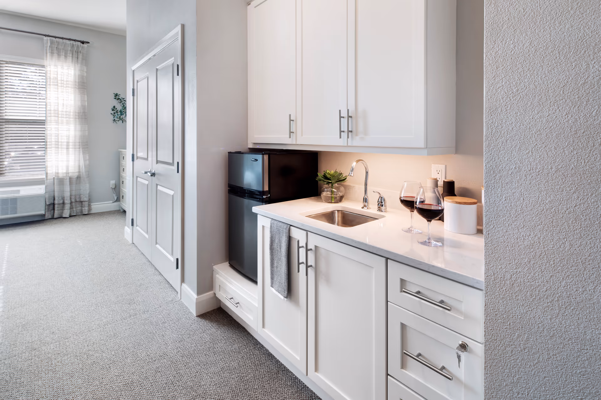 A small kitchenette area with white cabinets, a stainless steel sink, a mini refrigerator, and a countertop with two glasses of red wine and a small plant. The area is carpeted and adjacent to a window with sheer curtains and blinds.