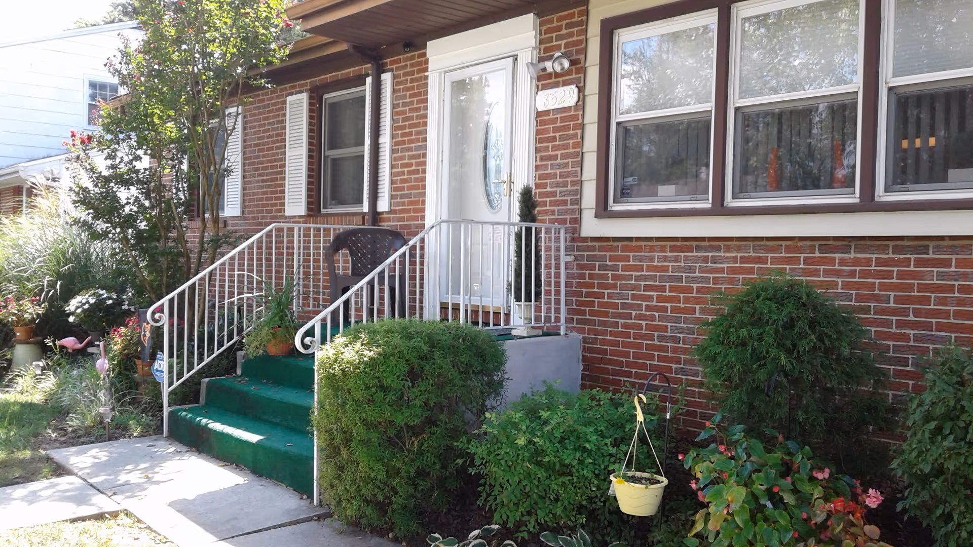 Front entrance of a brick house with green-carpeted steps, white metal railing, potted plants and shrubs.