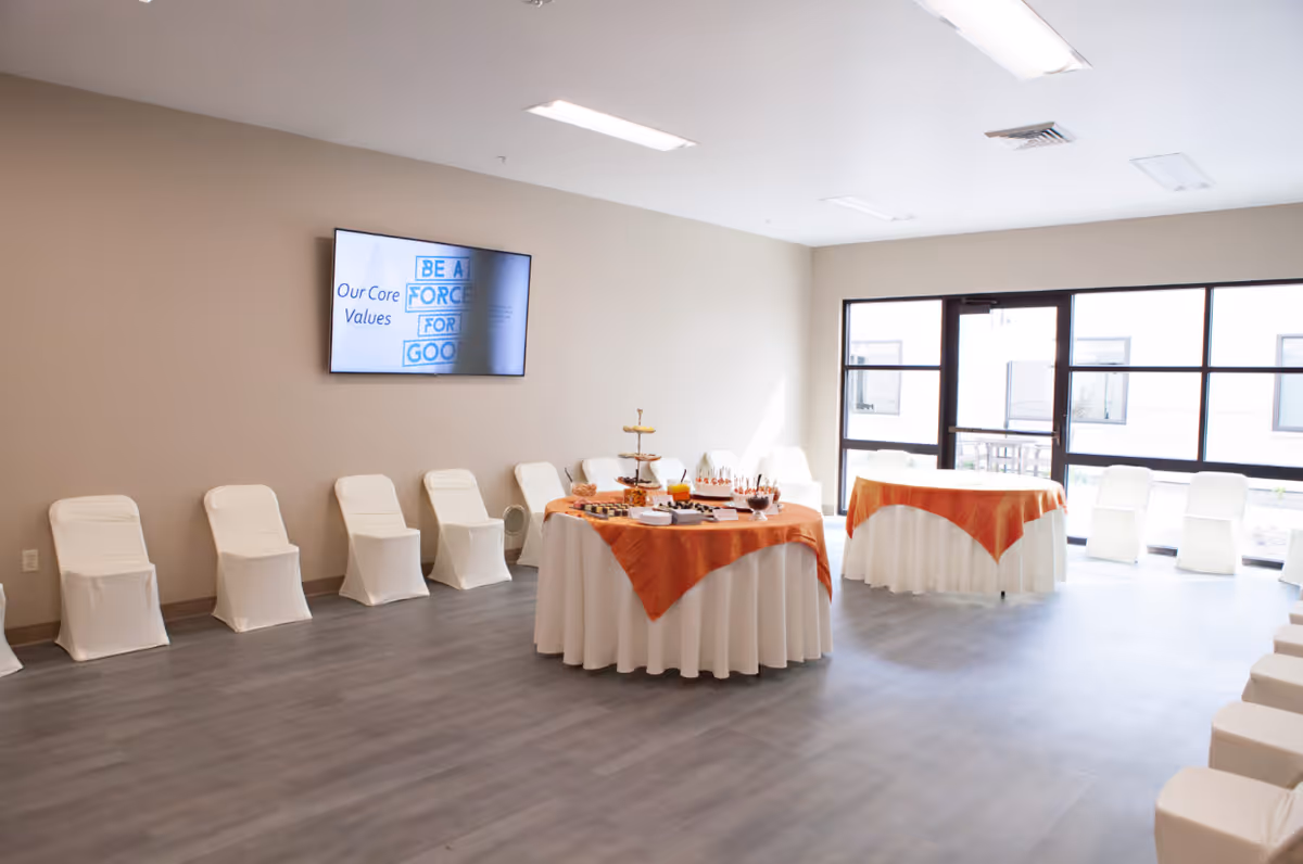 A bright, spacious room with white chairs covered in white fabric arranged along the walls. Two round tables with white tablecloths and orange overlays are set up in the center, displaying various snacks and desserts. A large window and glass door allow natural light to fill the room. A wall-mounted screen displays the text 'Our Core Values' and 'Be a Force for Good'.