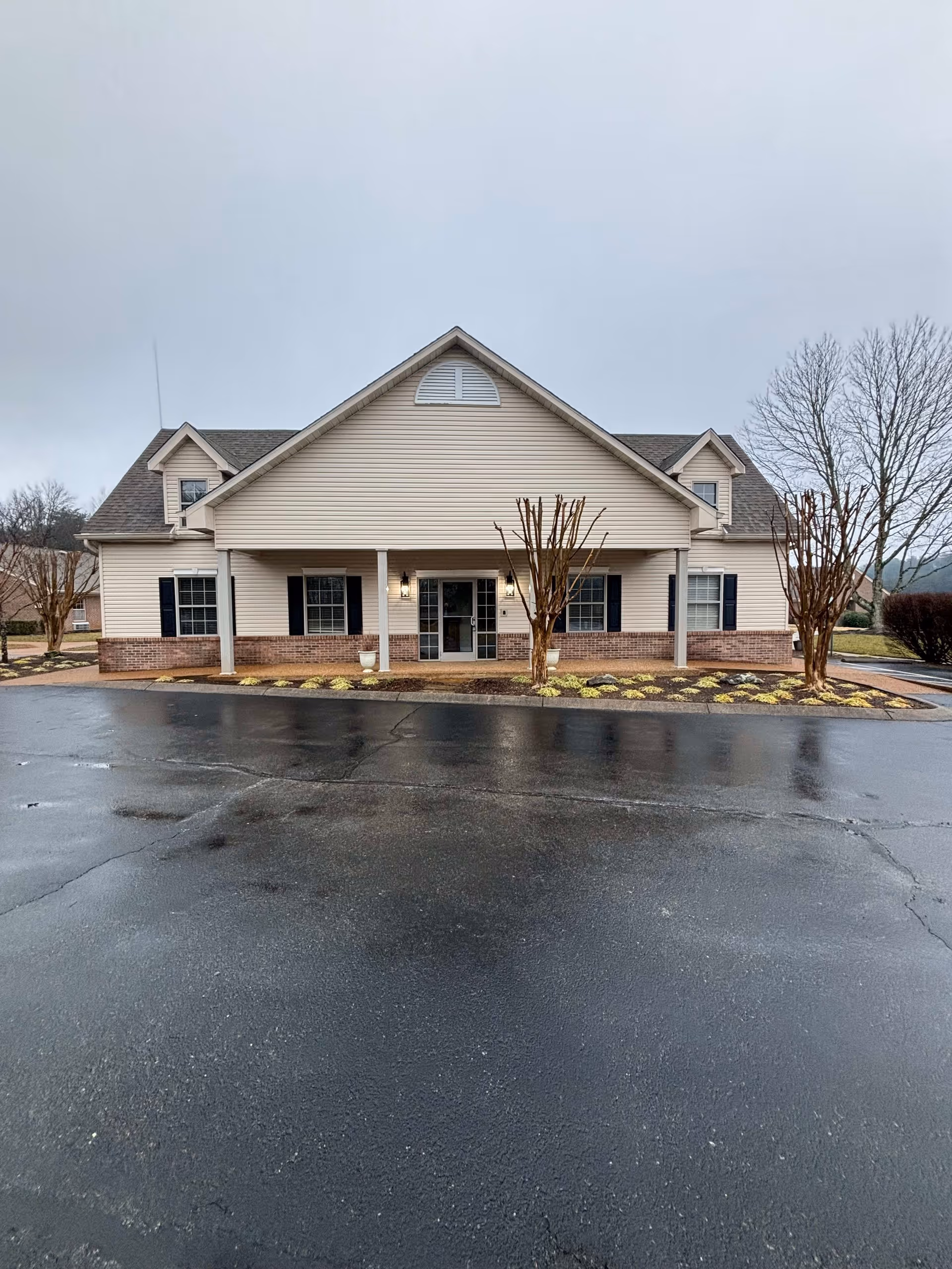 Front exterior view of a single-story senior living facility building with beige siding, brick accents, and a covered entrance supported by two columns. There are leafless trees and small shrubs in front of the building, and the ground is wet from recent rain under an overcast sky.