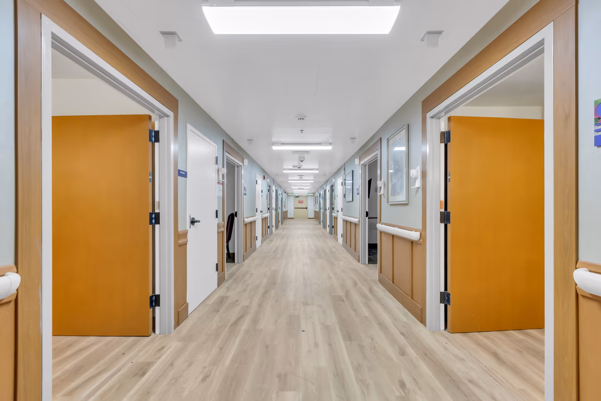 Long well-lit nursing facility hallway with open wooden doors, handrails, and light wood flooring.