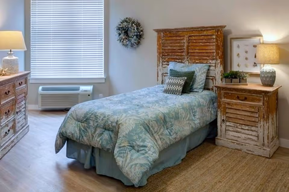 A cozy bedroom with a single bed featuring a blue and green floral comforter and matching pillows. The bed has a rustic wooden headboard. To the right of the bed is a distressed wooden nightstand with a lamp, a small plant, and framed artwork on the wall. On the left side of the room is a matching wooden dresser with a lamp and decorative items. A window with closed blinds is visible behind the bed, and the floor is wooden with a woven rug near the bed.