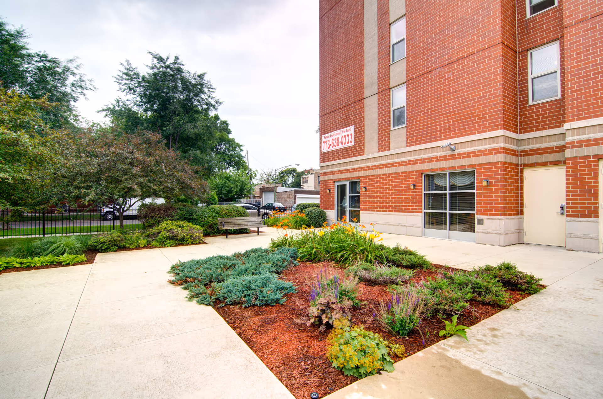 Outdoor view of a senior living facility with a landscaped garden area featuring various plants and flowers, a bench, and a brick building with windows and a door in the background.