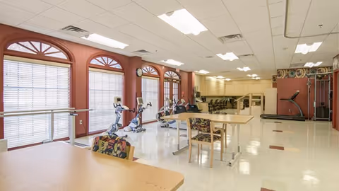 Large rehabilitation/exercise room with stationary bikes, a treadmill, parallel bars, and tables and chairs beneath arched windows.