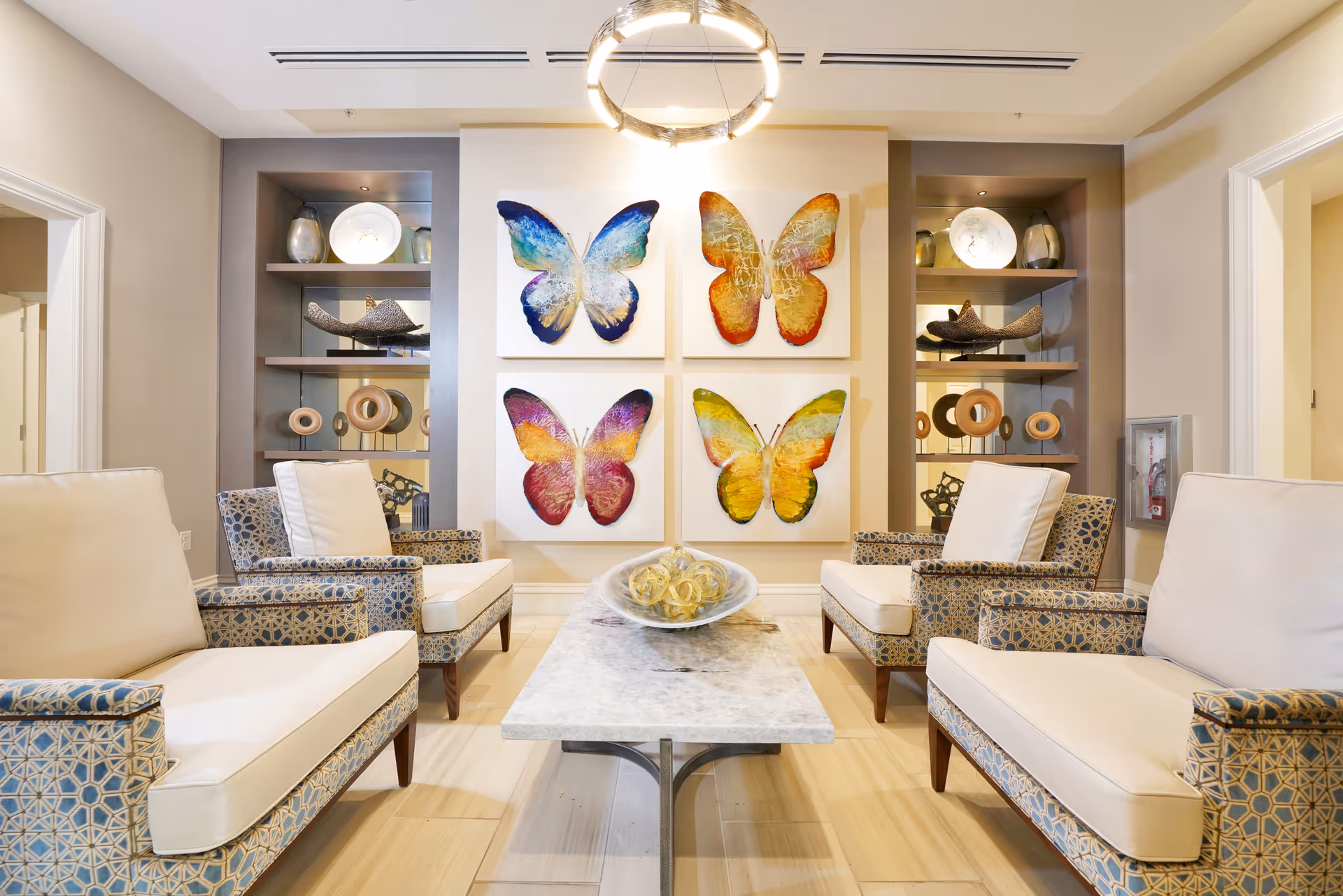 A bright and modern sitting area with four patterned armchairs featuring white cushions arranged around a rectangular marble coffee table. On the wall behind the chairs are four colorful butterfly paintings in blue, orange, purple, and yellow hues. Built-in shelves on either side of the paintings display decorative items including vases and sculptures. A circular chandelier hangs from the ceiling, illuminating the space.