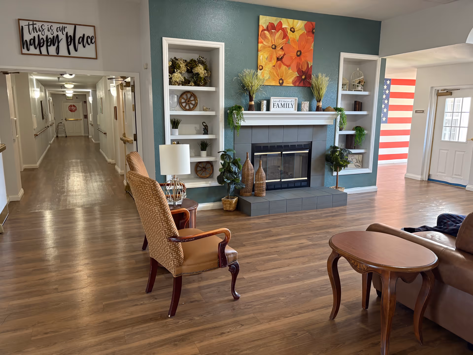 A cozy common area in a senior living facility featuring a fireplace with decorative plants and artwork above it. Two armchairs and a small wooden table are arranged on a wooden floor. A hallway extends to the left with handrails and a walker visible. A sign above the hallway reads 'this is our happy place'. To the right, an American flag mural is visible near a door with windows.