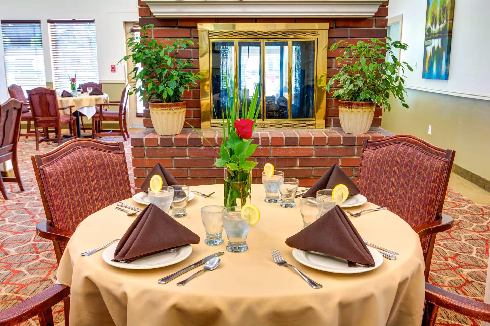 A round dining table set for four with brown napkins, water glasses garnished with lemon, and a rose centerpiece in front of a brick fireplace.