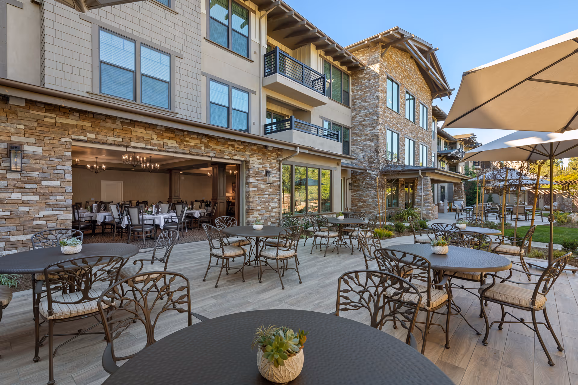 Outdoor patio area of The Vincent facility with multiple round metal tables and chairs featuring intricate back designs. Some tables have small potted plants. Large umbrellas provide shade. The patio is adjacent to a building with stone and beige siding, large windows, and an open dining room visible inside with tables and chairs set for meals.