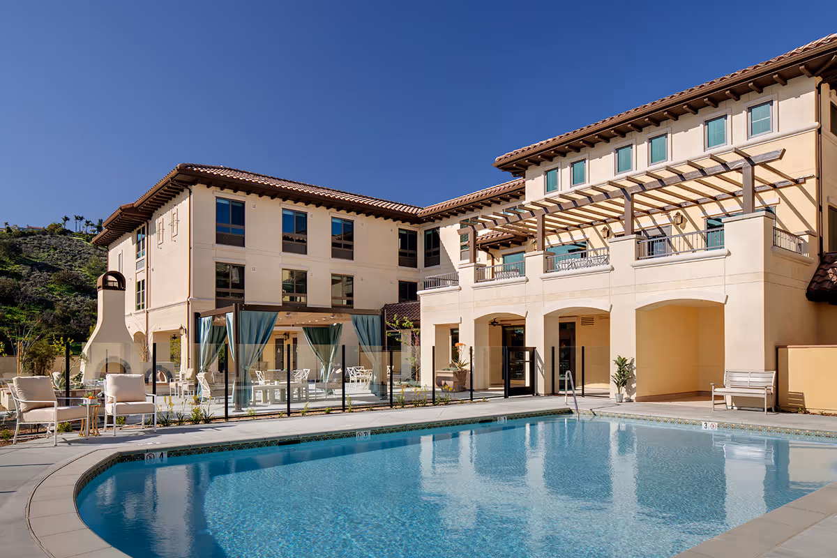 Outdoor pool and patio area with lounge chairs, cabanas, and a multi-story Mediterranean-style building under a clear blue sky.