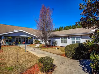 Outdoor view of a senior living facility with a paved walkway leading to a covered entrance. The building is single-story with white siding and multiple windows. There are bushes, a leafless tree, and a bench along the walkway under a clear blue sky.