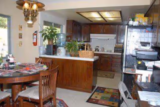 Interior view of a kitchen and dining area with wooden cabinets, a round dining table with chairs, a water dispenser, plants, and various kitchen appliances. The space has a cozy and homey atmosphere with a colorful rug on the floor and a chandelier above the dining table.