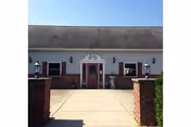 Front exterior of a single-story building with a centered red door, white siding, brick pillars flanking a paved entryway, and a clear blue sky.