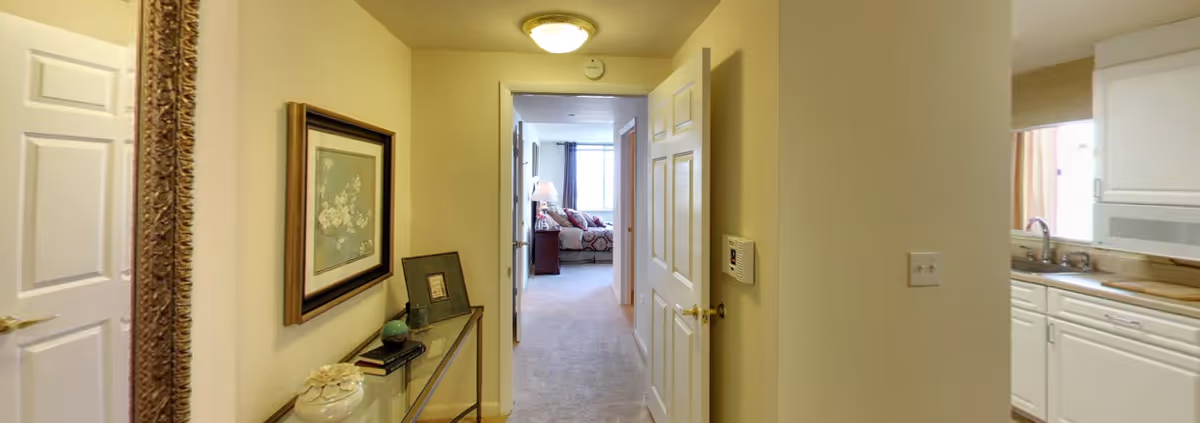 View down a hallway in a senior living residence with a glass console table holding decorative items and books on the left wall, a framed floral picture above it, and a doorway leading to a bedroom with a bed and nightstand visible in the background. To the right, there is a partial view of a kitchen with white cabinets, a sink, and a microwave.